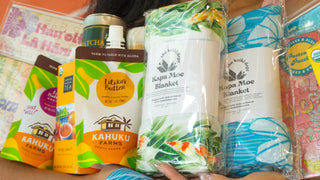 A person holds multiple packaged Kaukuku products, including blankets and snack pouches, against a bright outdoor background. Visible products include a white Kapa Moe Blanket, a blue patterned blanket, a yellow Mac-Nut Crumble pouch, and colorful snack boxes.